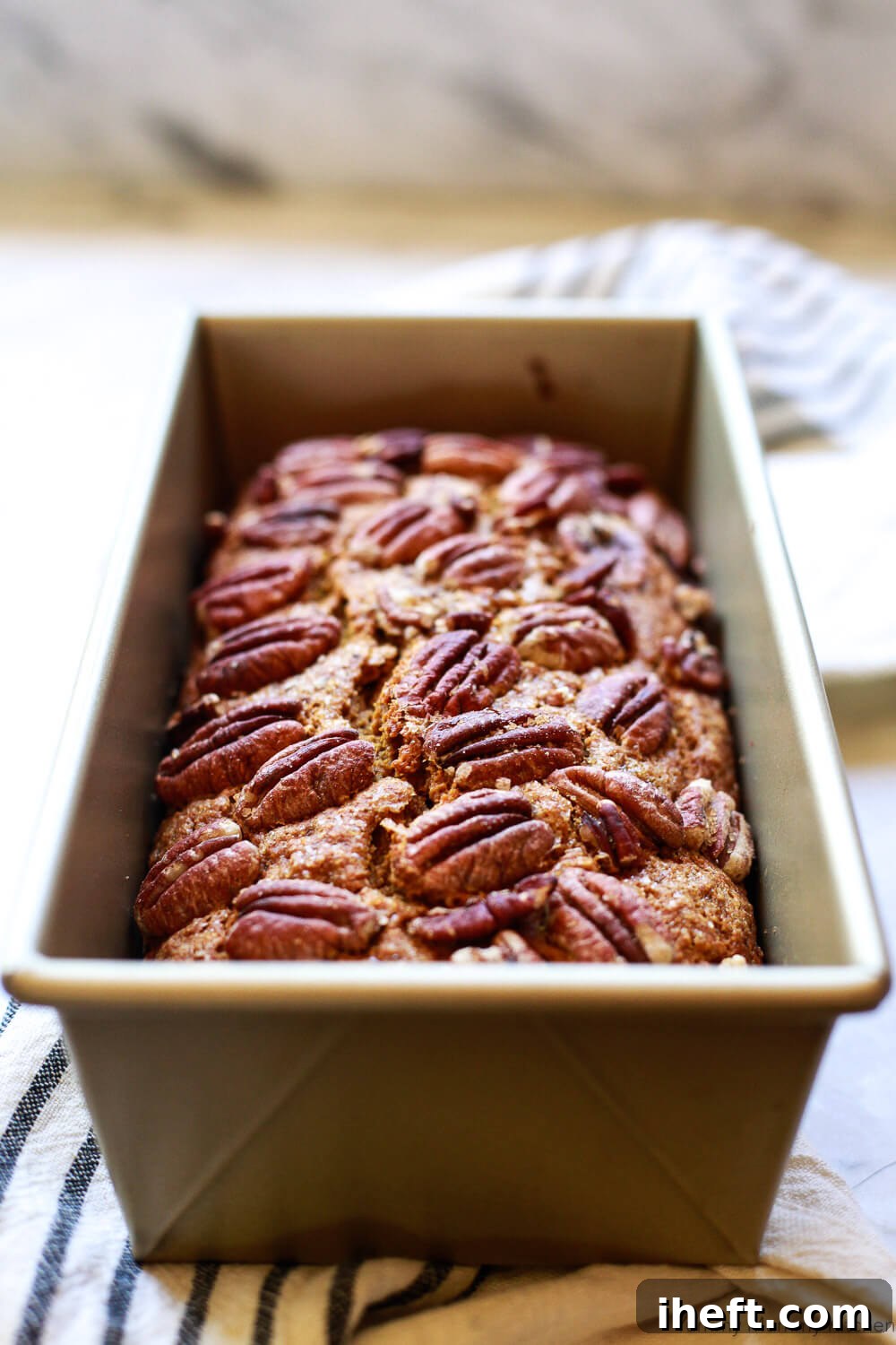 A gold loaf pan filled with vegan pumpkin bread topped with whole pecans on a marble countertop with a black and white striped towel. 