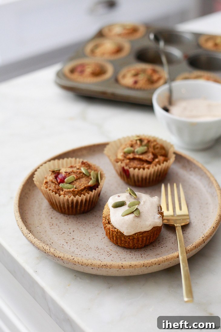 Gluten free vegan pumpkin muffins on a ceramic plate. One is topped with cream cheese frosting and pepitas. A fork sits on the plate and muffin tin in the background in a white kitchen. 