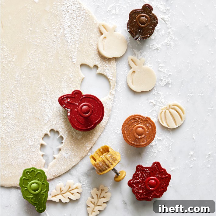 A close-up of various fall-themed pastry cutters, shaped like leaves and pumpkins, pressing designs into a rolled-out sheet of pie crust dough.