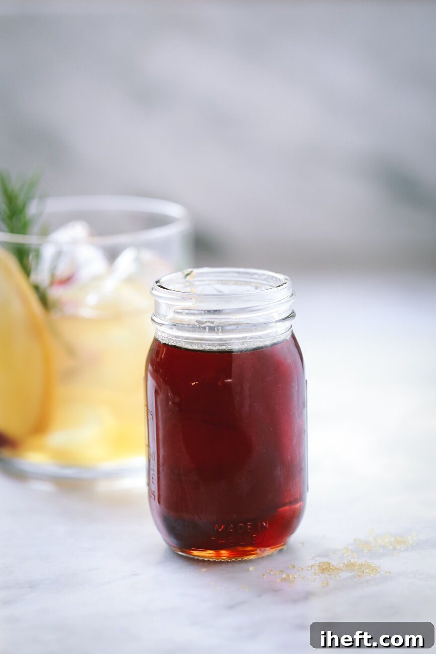 A small jar of homemade demerara syrup on a white countertop with an old fashioned cocktail in the background.