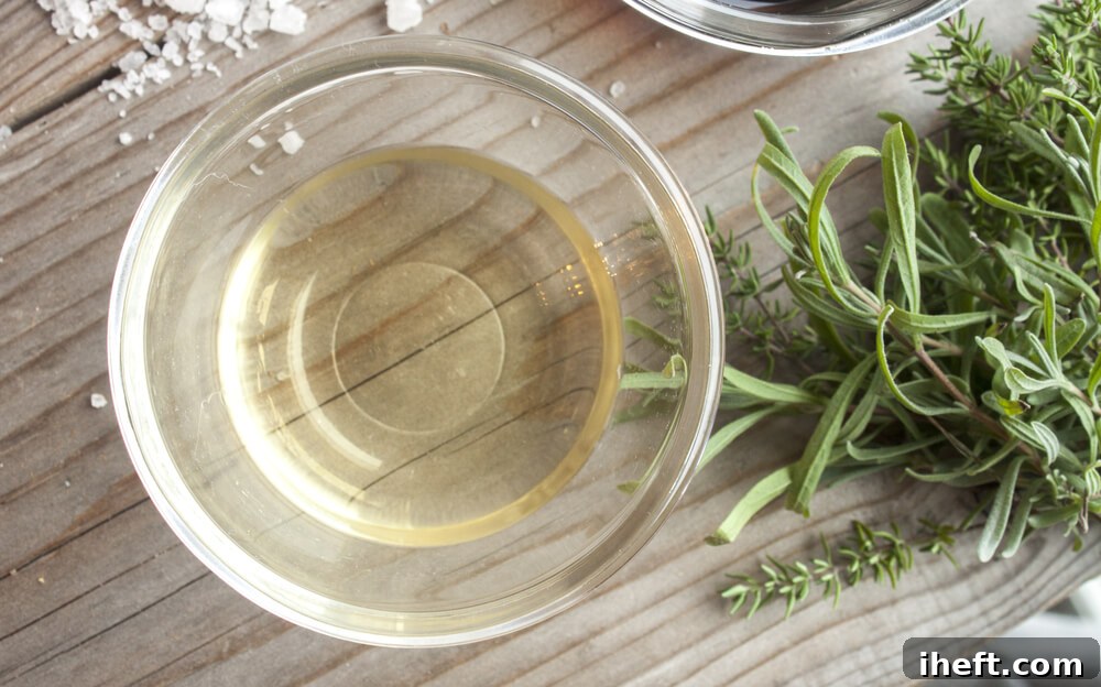A small, clear glass bowl filled with white wine vinegar rests on a rustic wooden cutting board, accompanied by fresh green herbs like rosemary and thyme, suggesting its use in culinary preparations.