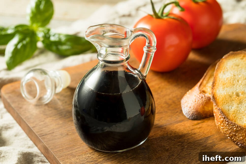 A small, elegant glass dressing bottle filled with rich, dark balsamic vinegar is placed on a rustic wooden cutting board, surrounded by fresh tomatoes and toasted bread slices, suggesting its use in appetizers or salads.