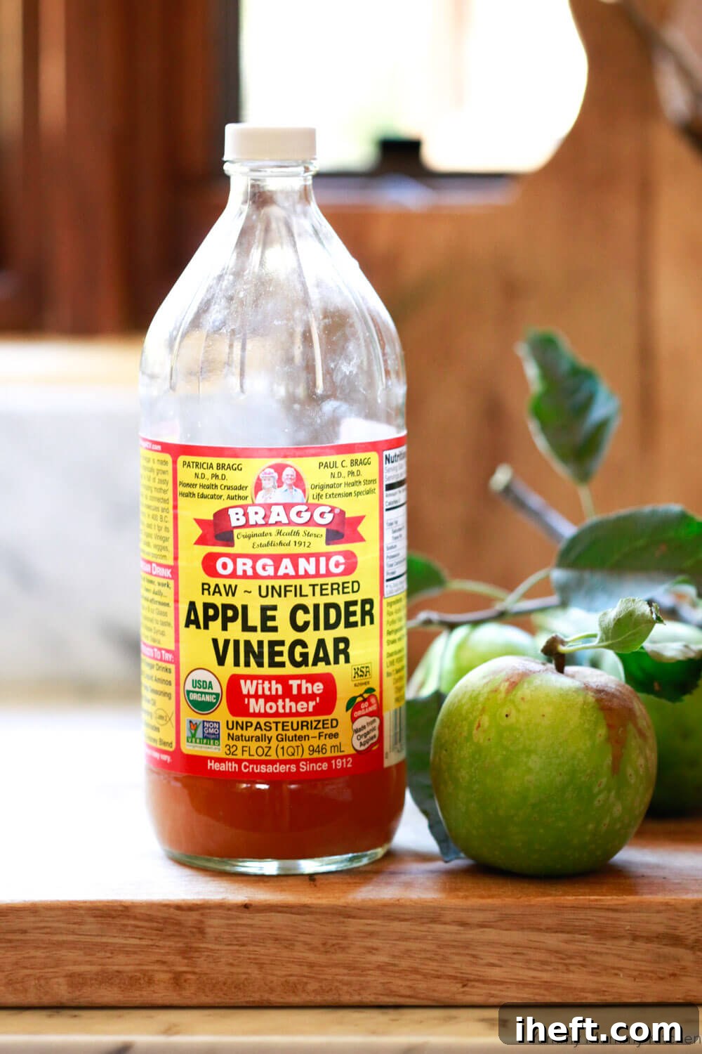 A bottle of Bragg apple cider vinegar with its label prominently displayed stands on a natural wooden cutting board, surrounded by fresh, vibrant red apples, highlighting its origin and natural qualities.