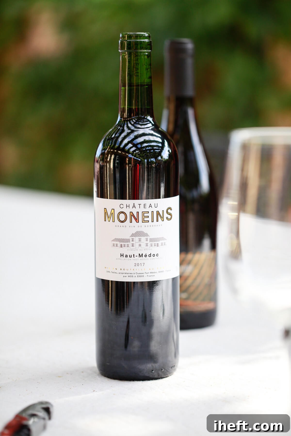 A bottle of elegant red wine stands upright on a rustic outdoor wooden table, suggesting a relaxed evening gathering or a culinary preparation. The natural lighting highlights the rich color of the wine.