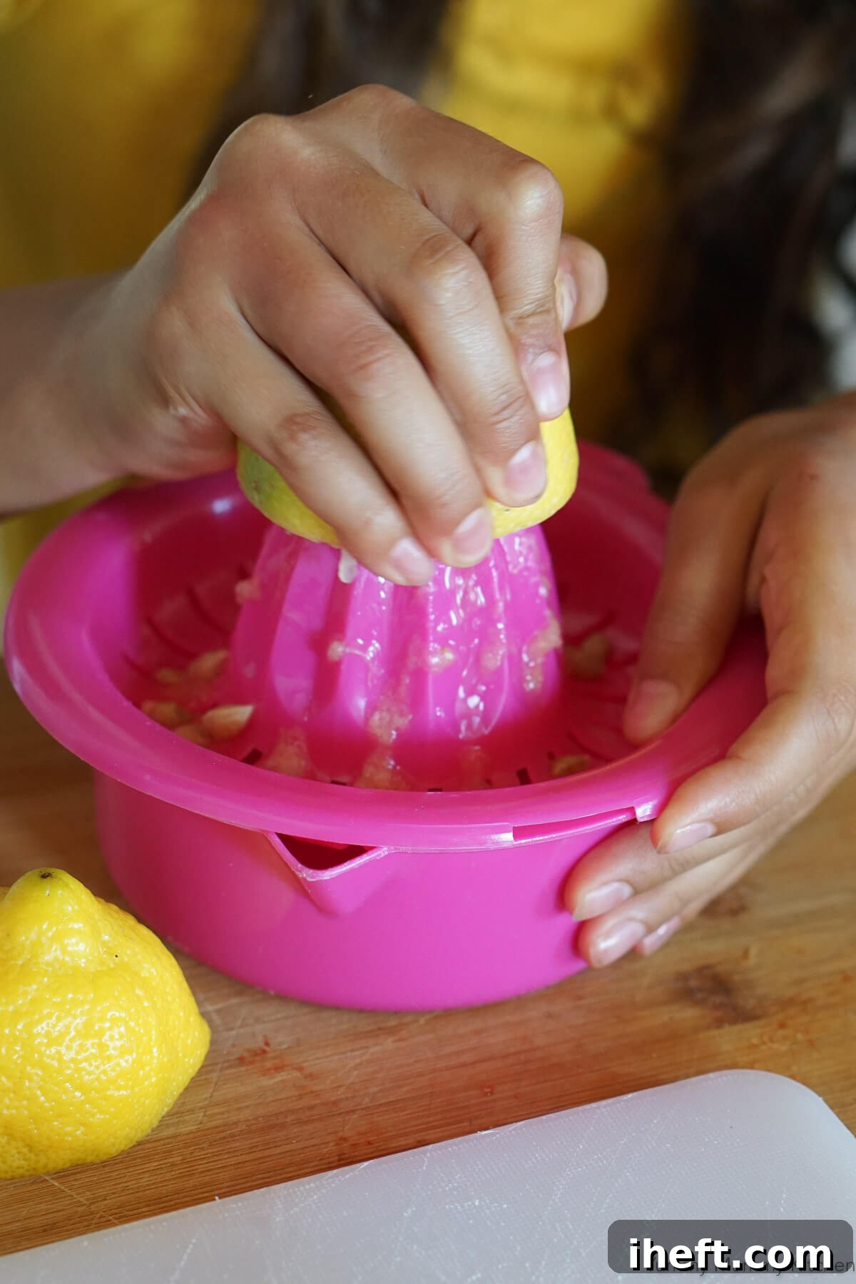 A young child's hands are diligently juicing a vibrant yellow lemon on a bright pink citrus juicer, creating a refreshing splash of lemon juice.