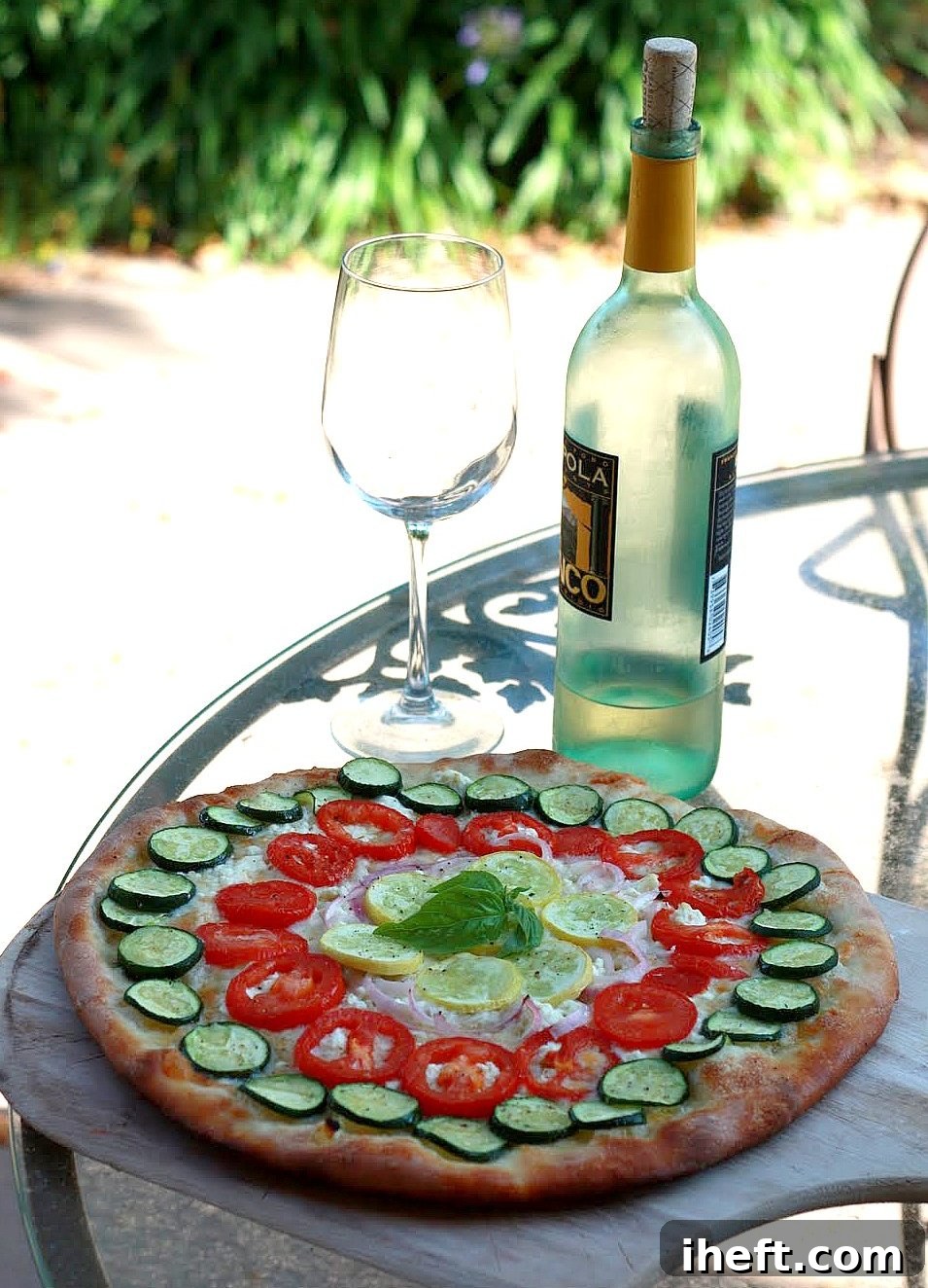 A vibrant rainbow vegetable pizza, generously topped with thinly sliced zucchini, bright red tomatoes, sweet summer squash, and delicate purple onions, sits invitingly on a rustic patio table next to a bottle of crisp white wine. The scene exudes a warm, inviting atmosphere, perfect for a relaxed outdoor meal.