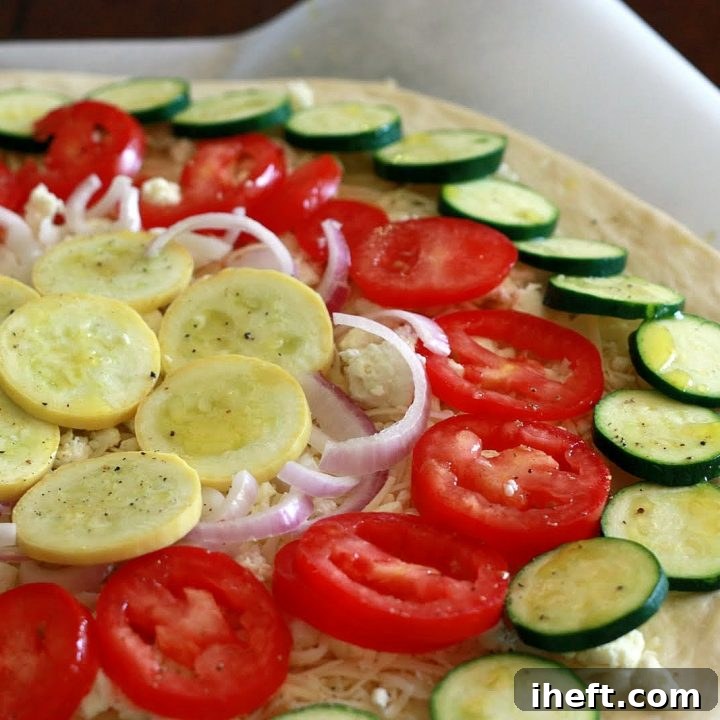 A vibrant rainbow vegetable pizza, generously topped with thinly sliced zucchini, bright red tomatoes, sweet summer squash, and delicate purple onions, sits invitingly on a rustic patio table next to a bottle of crisp white wine. The scene exudes a warm, inviting atmosphere, perfect for a relaxed outdoor meal.