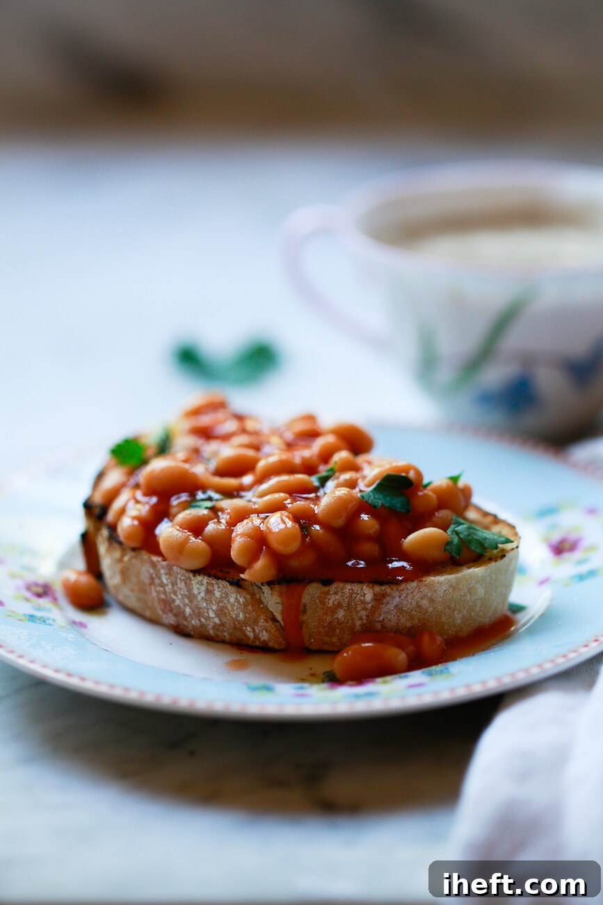 A slice of British beans on toast on a small plate on a marble kitchen countertop. The beans are garnished with parsley and a small teacup sits in the background. 