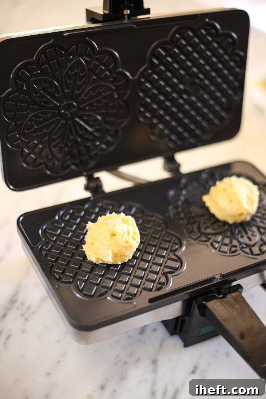 Two neatly scooped balls of pizzelle dough are placed onto the heated plates of a pizzelle press, just before the lid is closed for cooking.