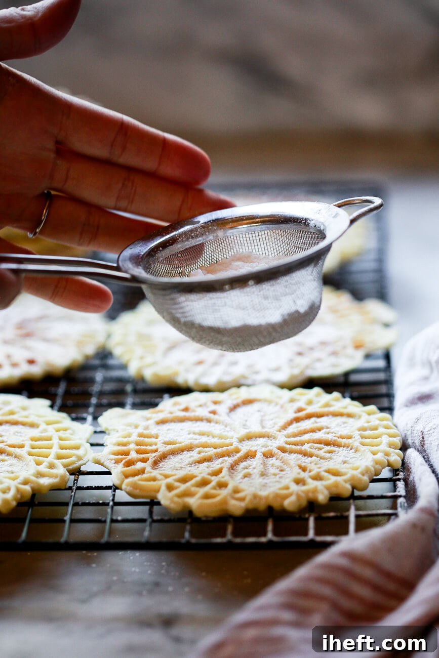 Powdered sugar is delicately sifted over a stack of freshly baked pizzelle cookies through a sifter, adding a beautiful finish.