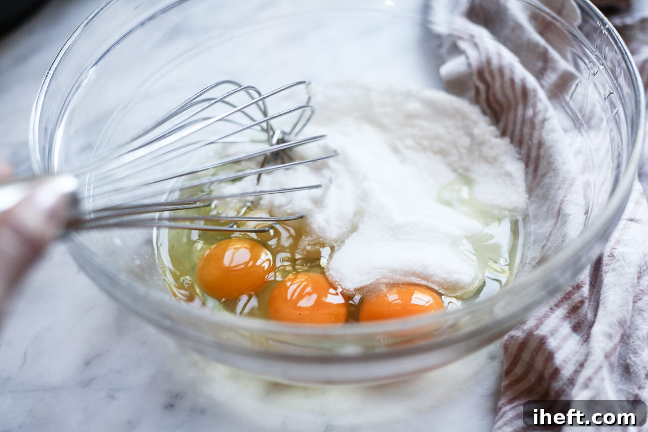 Three large eggs and granulated sugar are whisked together in a clear glass bowl, creating a creamy mixture.