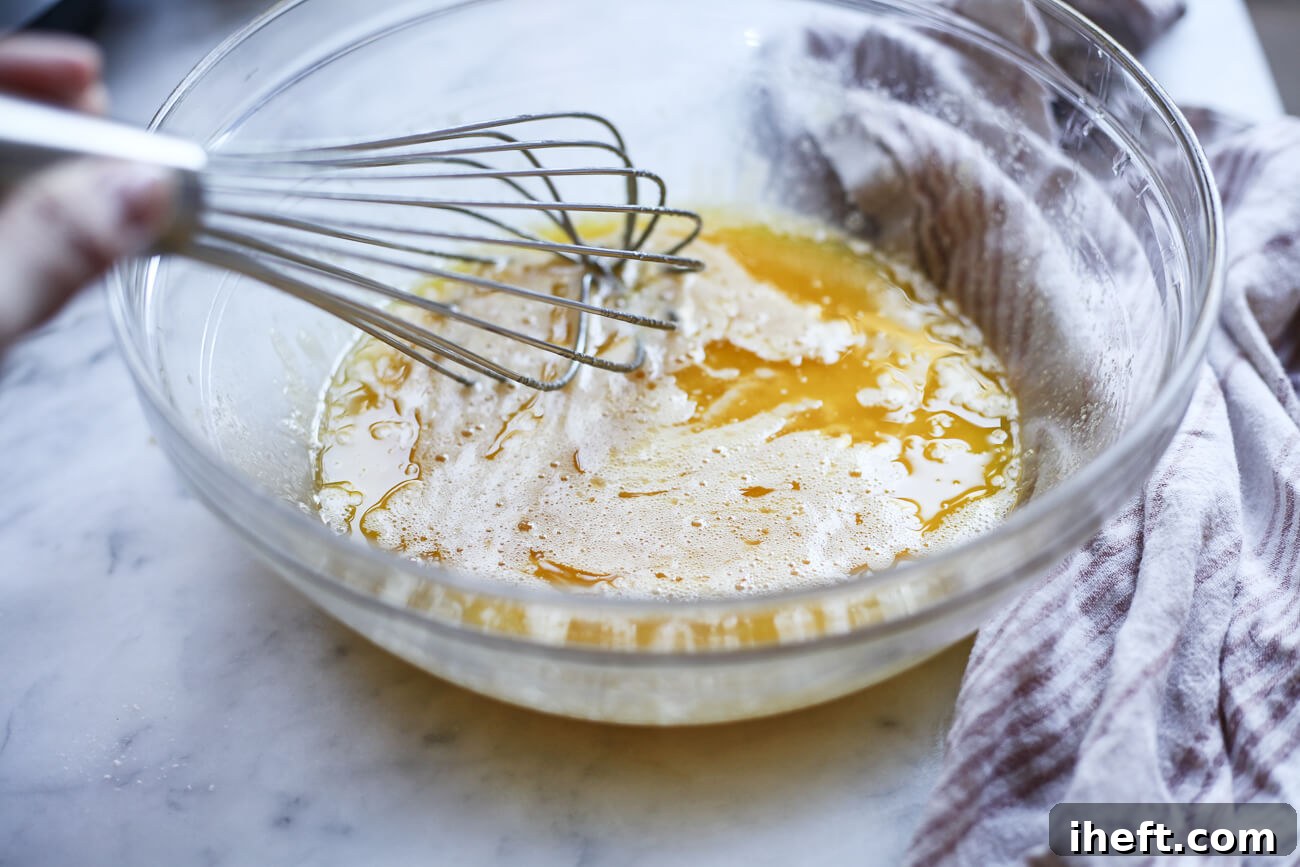 Melted butter is being whisked into a creamy mixture of eggs and sugar in a glass bowl.