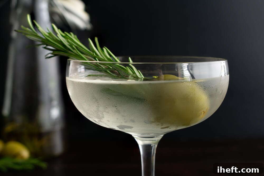 A close-up photo of a perfectly chilled dirty martini in a sleek coupe glass, garnished with a fresh sprig of rosemary and a vibrant green olive, ready to be enjoyed.