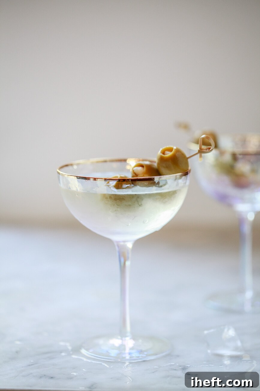 A beautiful Anthropologie coupe glass filled with a cold dirty martini, meticulously garnished with skewered garlic olives, set against the backdrop of a pristine white kitchen.