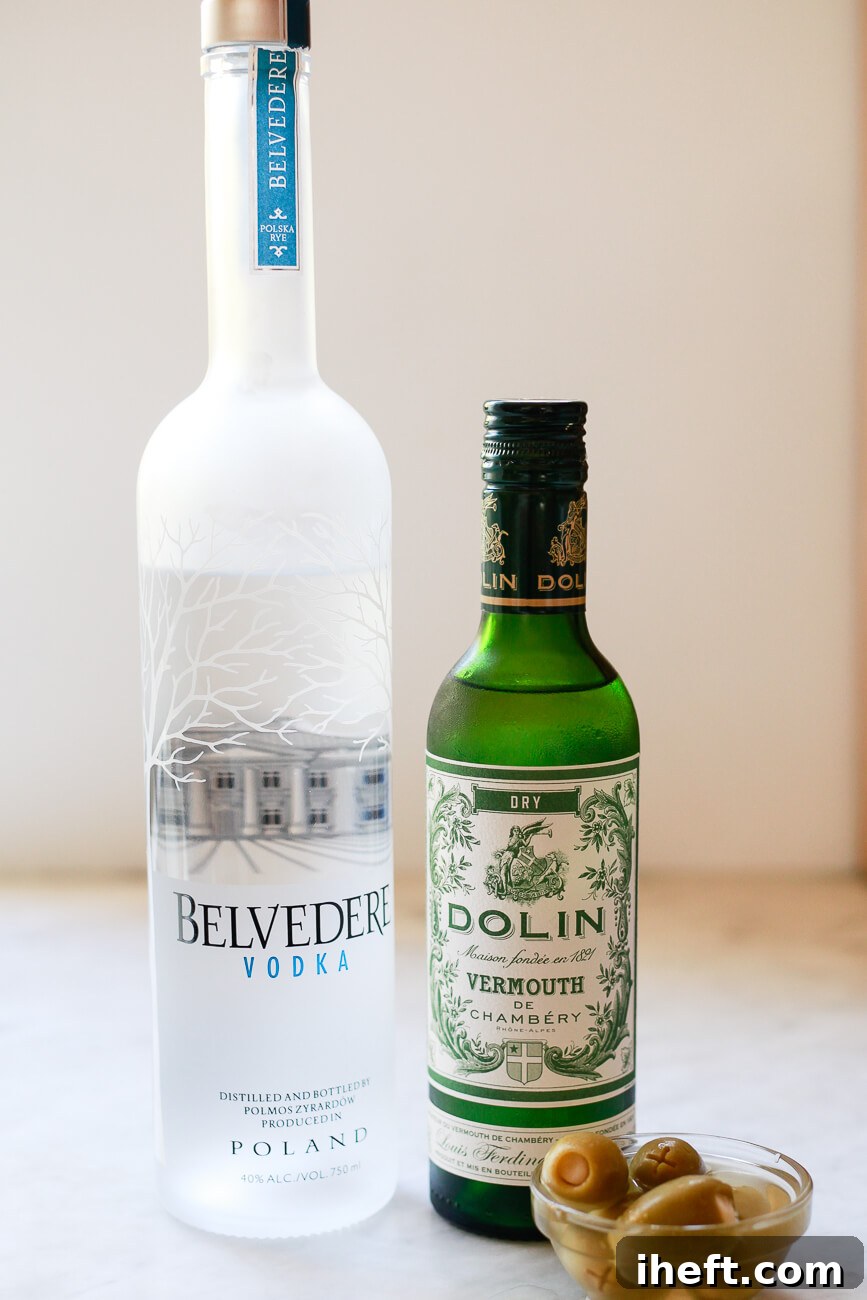 Bottles of premium vodka and dry white vermouth sit elegantly on a polished marble countertop, accompanied by a small dish of vibrant green olives – the essential components for a classic martini.