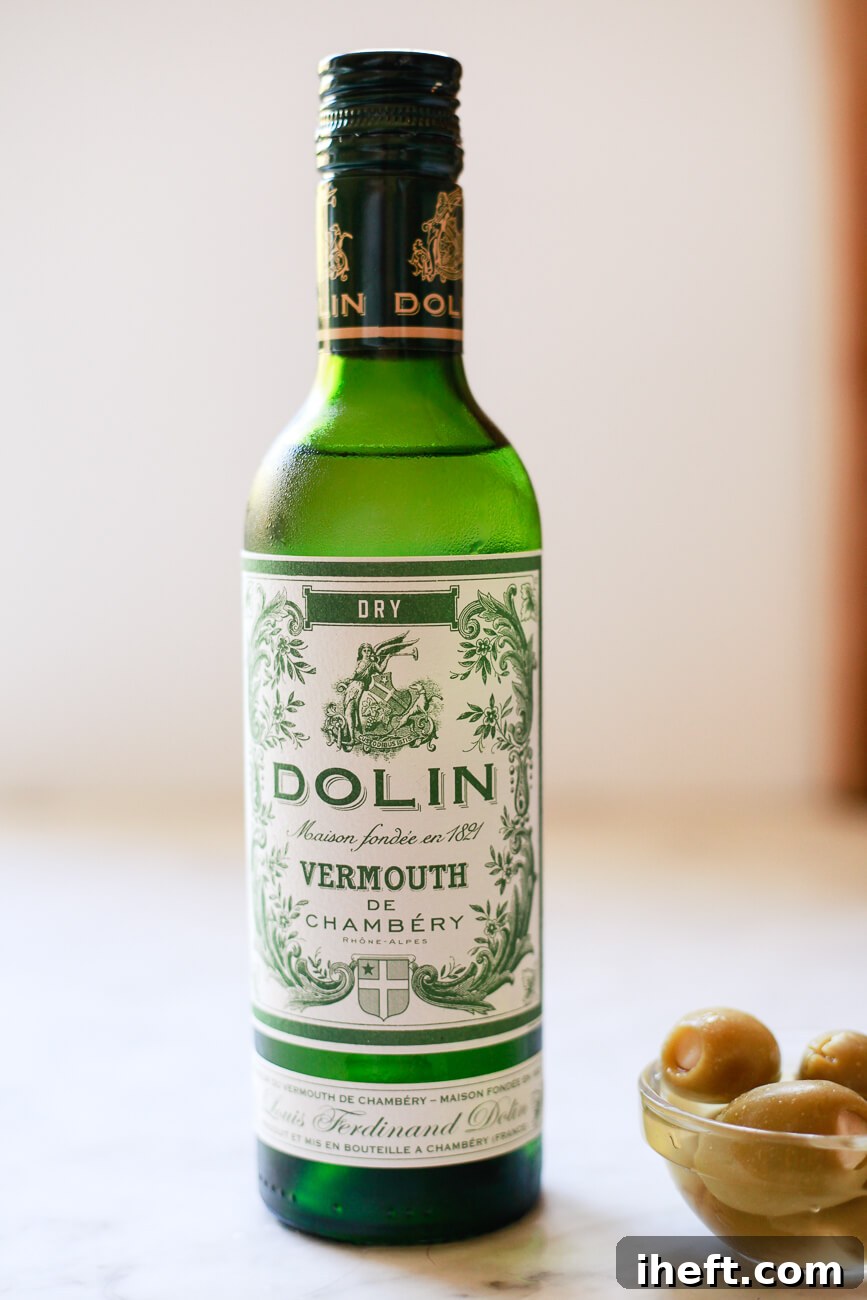 A small, elegant green bottle of Dolin dry white vermouth rests on a cool marble countertop, poised beside a ceramic dish overflowing with glistening green olives – the perfect martini pairing.