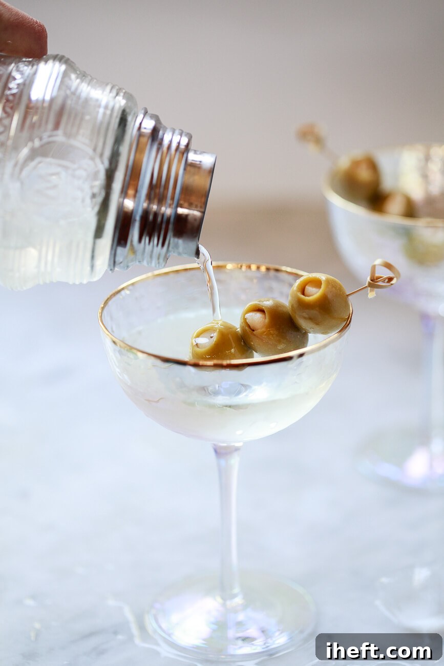 A vintage crystal cocktail shaker gracefully pours a freshly made homemade dirty martini into a sparkling coupe glass, elegantly garnished with skewered green olives.