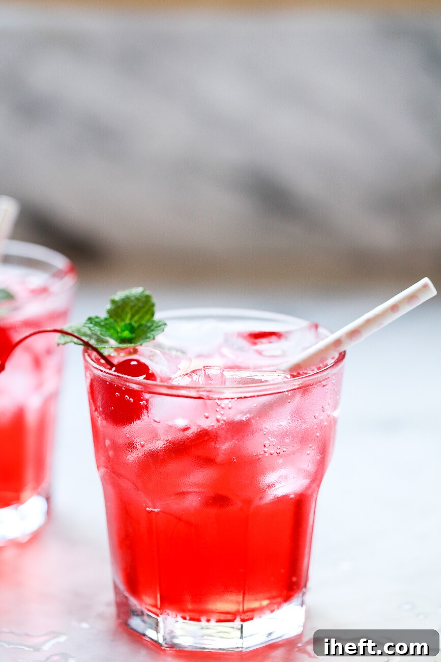 A close-up of a vibrant Shirley Temple drink, garnished with a bright red maraschino cherry and a fresh sprig of mint, served with a small straw on a sleek marble kitchen counter. The drink's pink hue is inviting and refreshing.