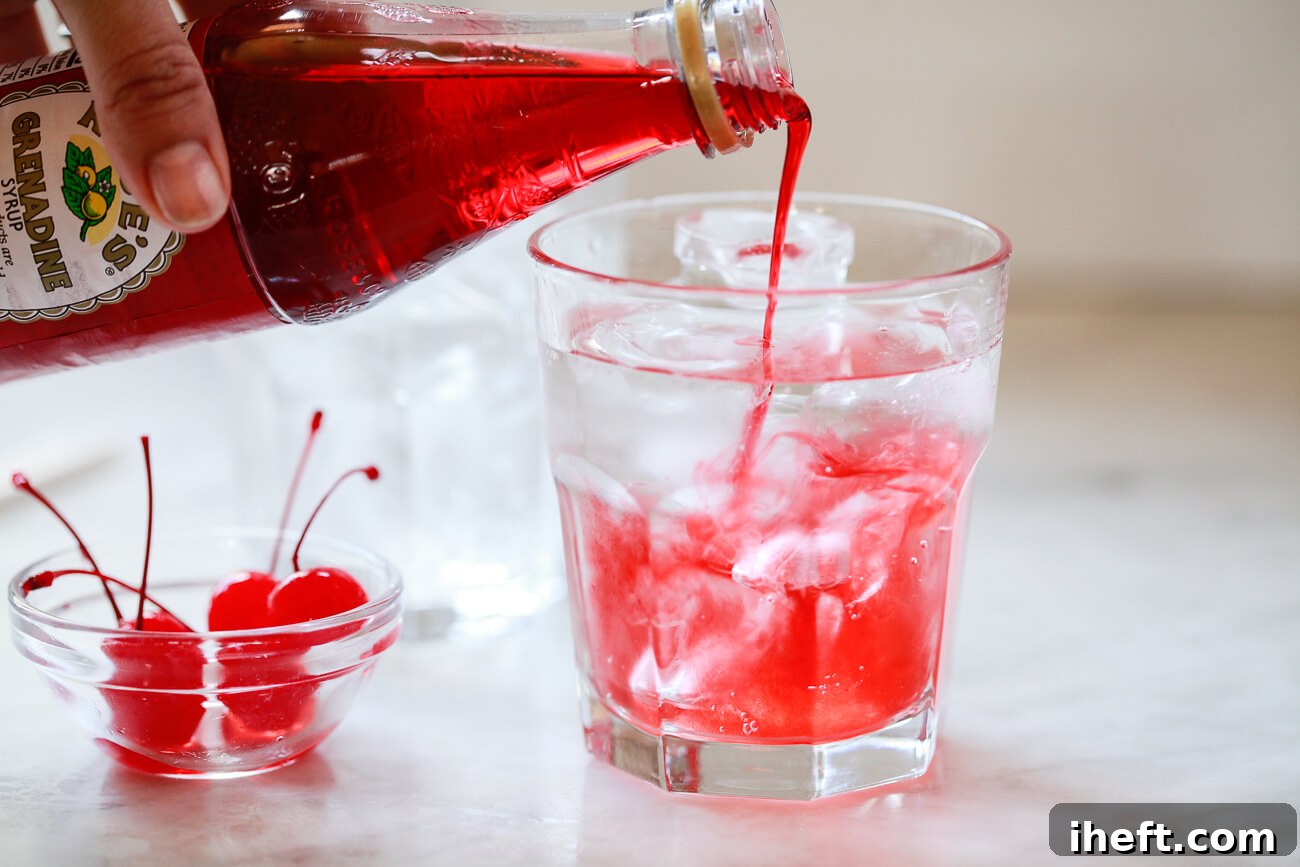 Rose's grenadine syrup being poured into a tall glass filled with clear lemon-lime soda, sitting on a marble counter. A small bowl of glistening maraschino cherries is placed beside it, ready for garnish.