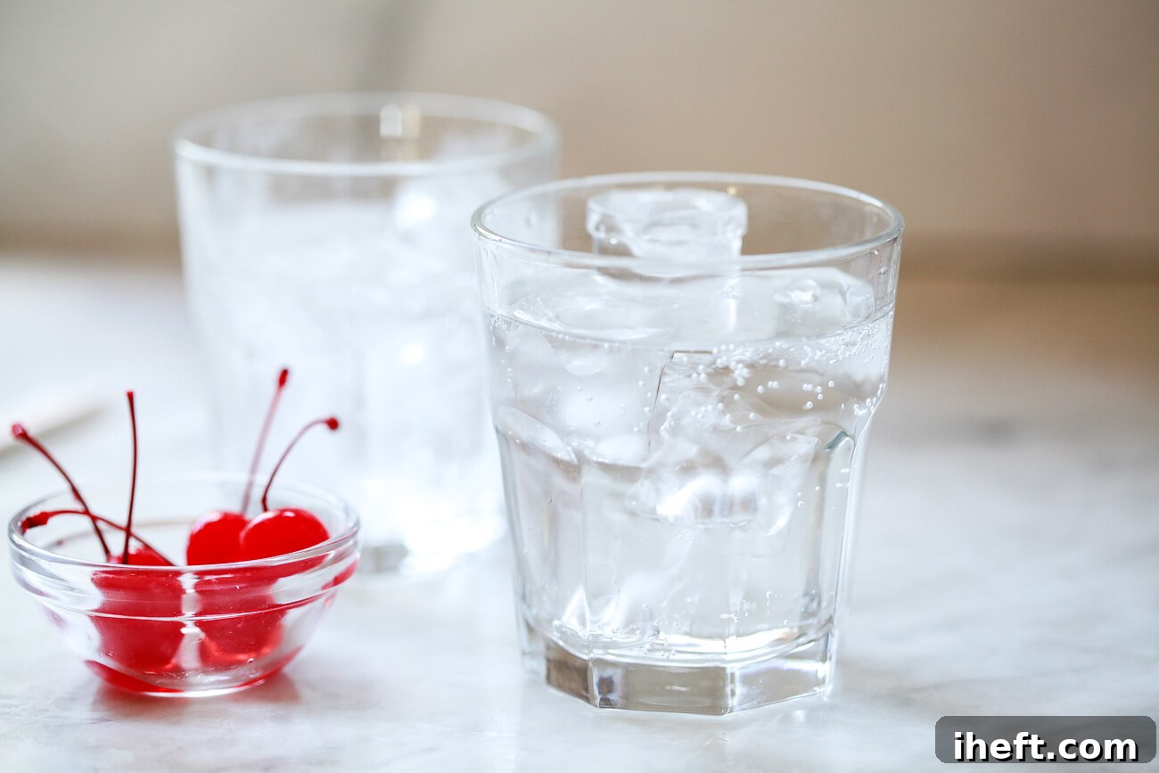 Two clear glasses are filled with ice and sparkling lemon-lime soda, ready for the next steps in making a Shirley Temple. A bowl of maraschino cherries is nearby.
