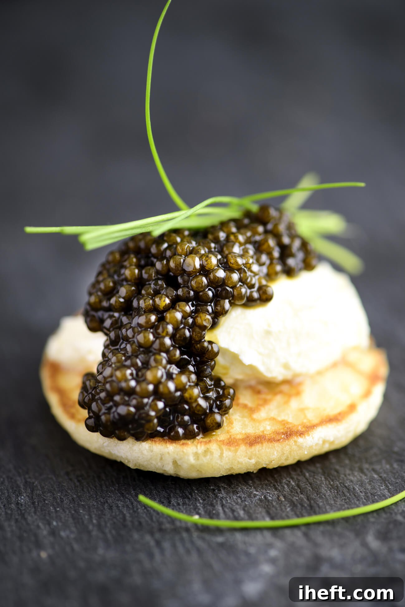 A close-up photo of a blini topped with creme fraiche, black caviar, and chives on a slate background. 