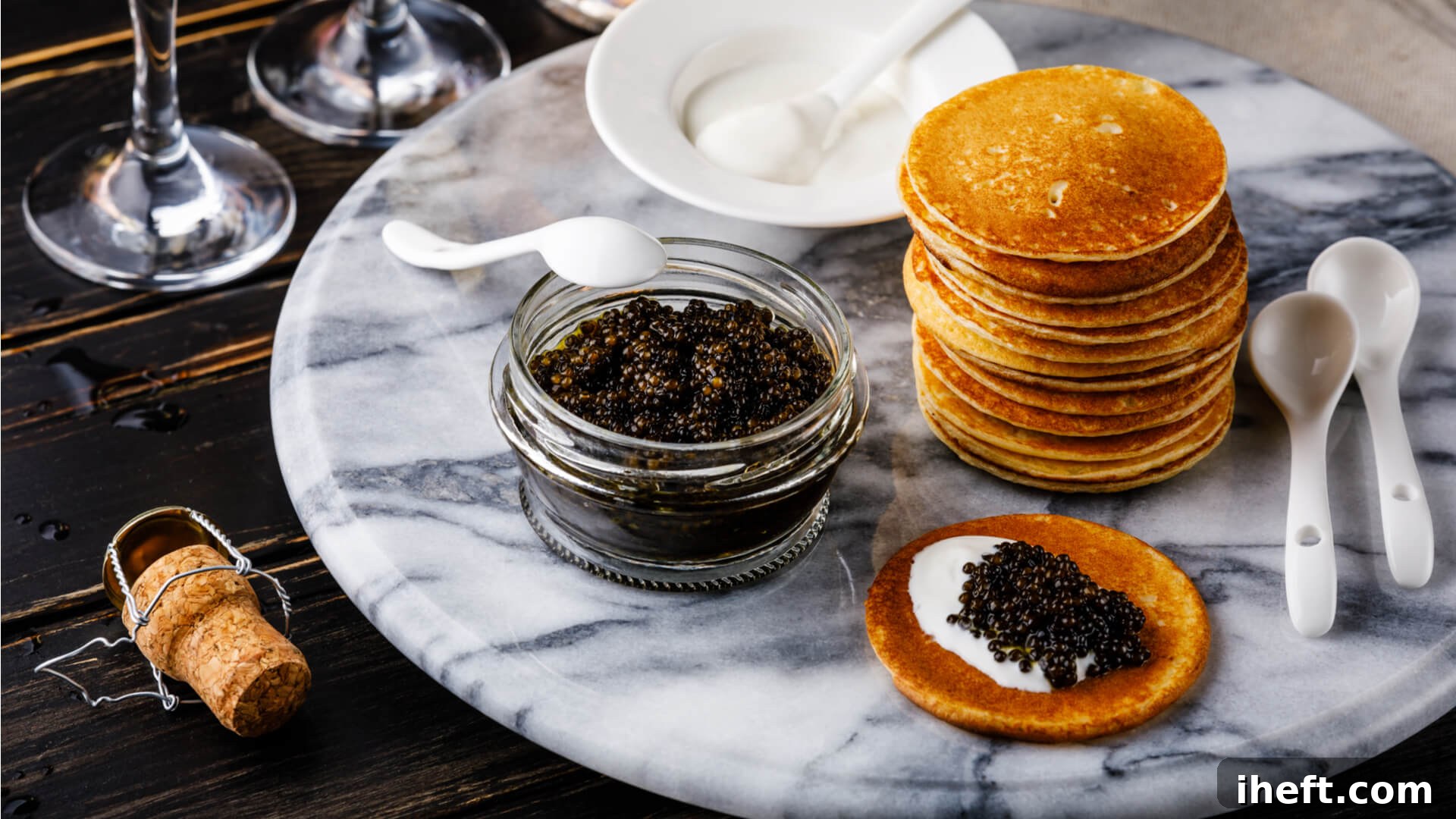 A glass bowl filled with black caviar and a white spoon sits on a marble platter next to blini Russian pancakes. 