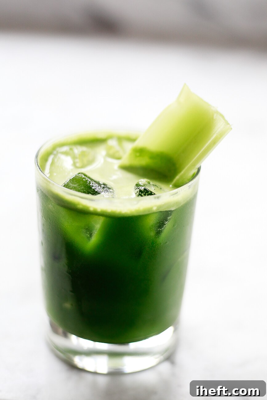 A pristine glass filled with homemade green detox juice and ice sits elegantly on a smooth marble countertop. The refreshing juice is artfully garnished with a crisp celery stick, ready to be enjoyed.