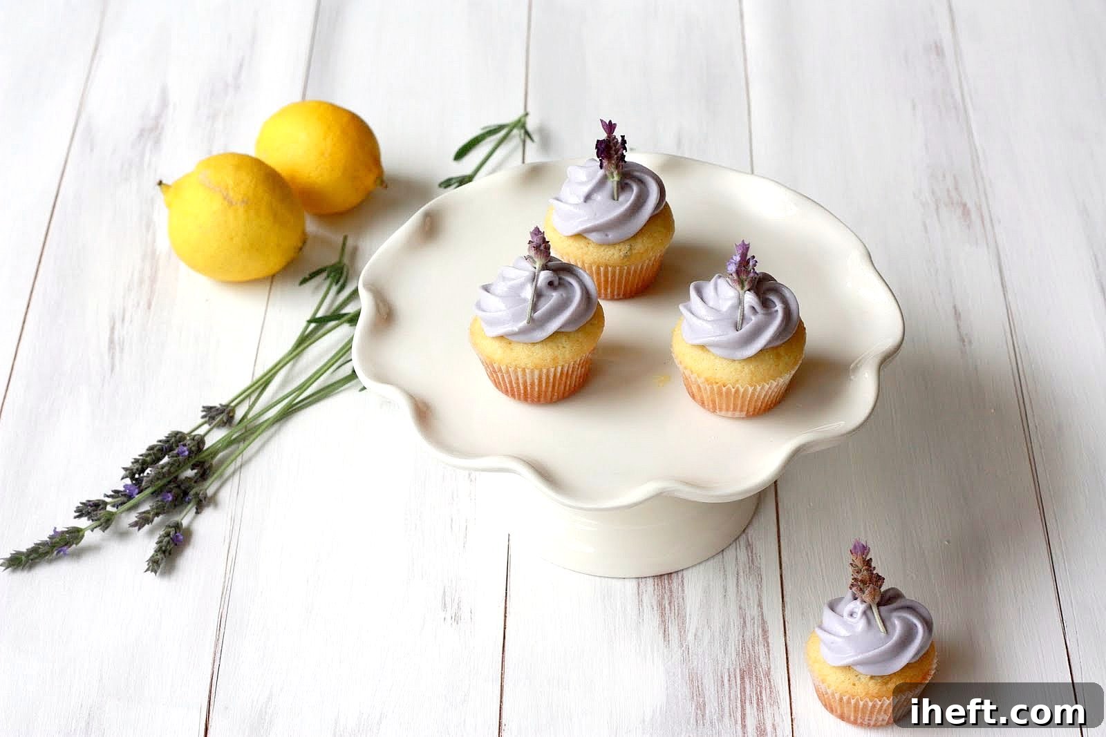 4 beautiful lavender cupcakes on a small cake stand with lemons and fresh lavender in the background. The cupcakes are topped with a single lavender flower.