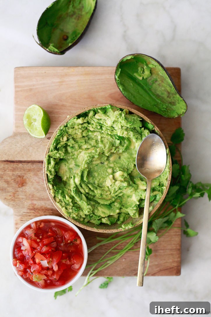 A bowl filled with homemade guacamole on a cutting board next to avocado peels and a cup of salsa. 
