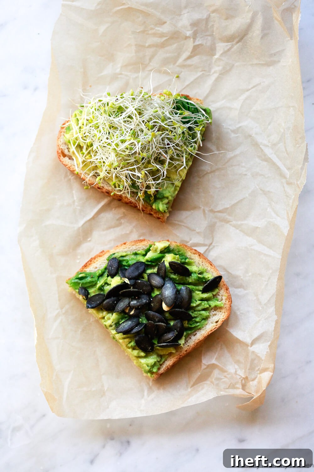 Two pieces of avocado toast on sourdough bread on a piece of parchment paper. One is topped with sprouts with the other is topped with pumpkin seeds. 