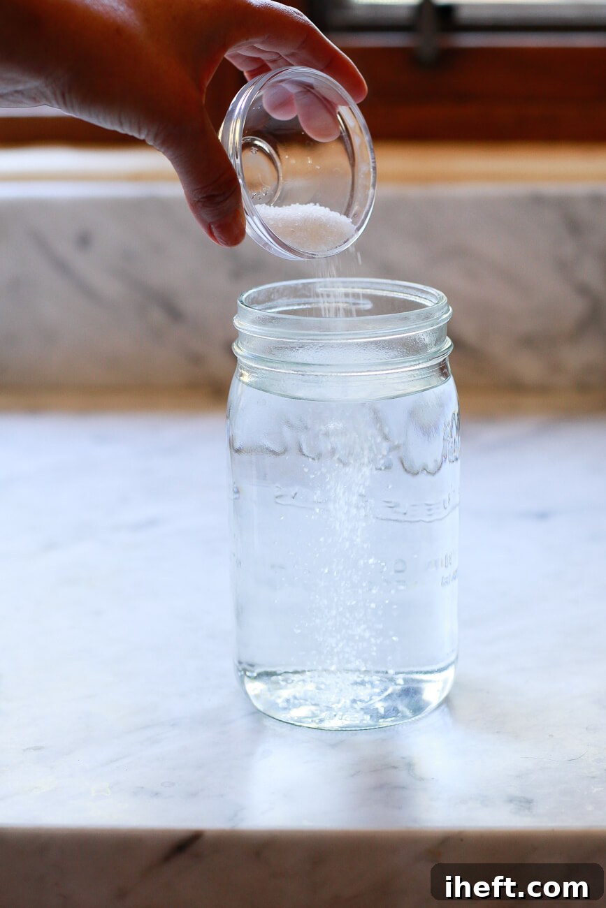 A hand pours a small dish of salt into a liter sized jar of warm water on a marble kitchen counter.