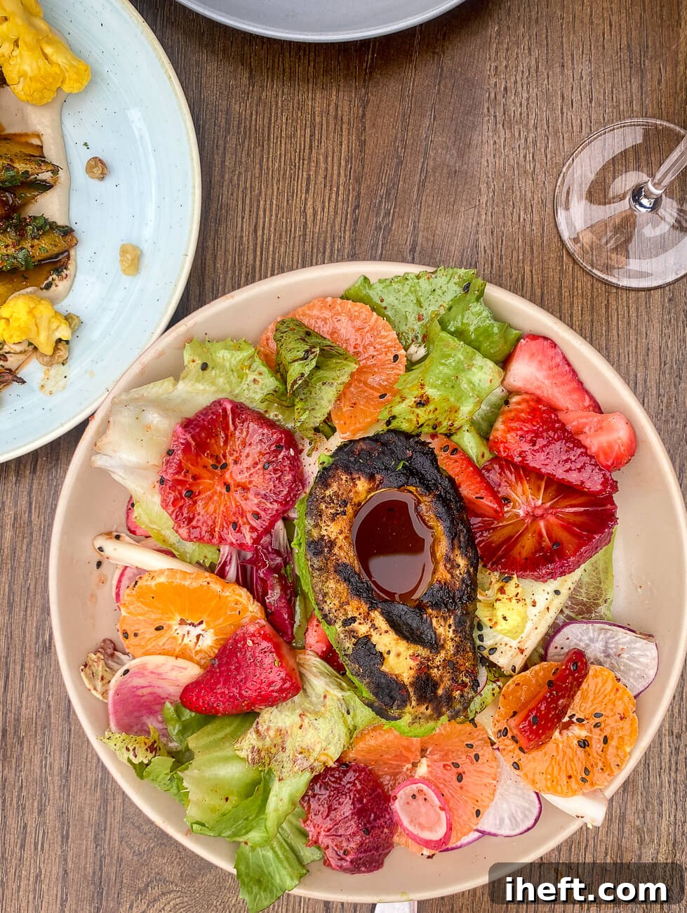 A vibrant, colorful salad composed of crisp Romaine lettuce, bright blood oranges, fresh strawberries, delicate radish slices, and a perfectly grilled avocado, all artfully arranged on a rustic wooden table.