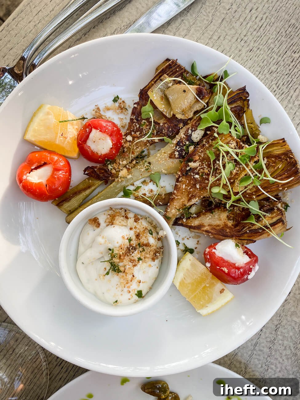 A plate of perfectly roasted Roman artichoke heart halves, elegantly topped with delicate microgreens and a sprinkle of breadcrumbs, served alongside a small dish of creamy aioli and vibrant peppadew peppers.