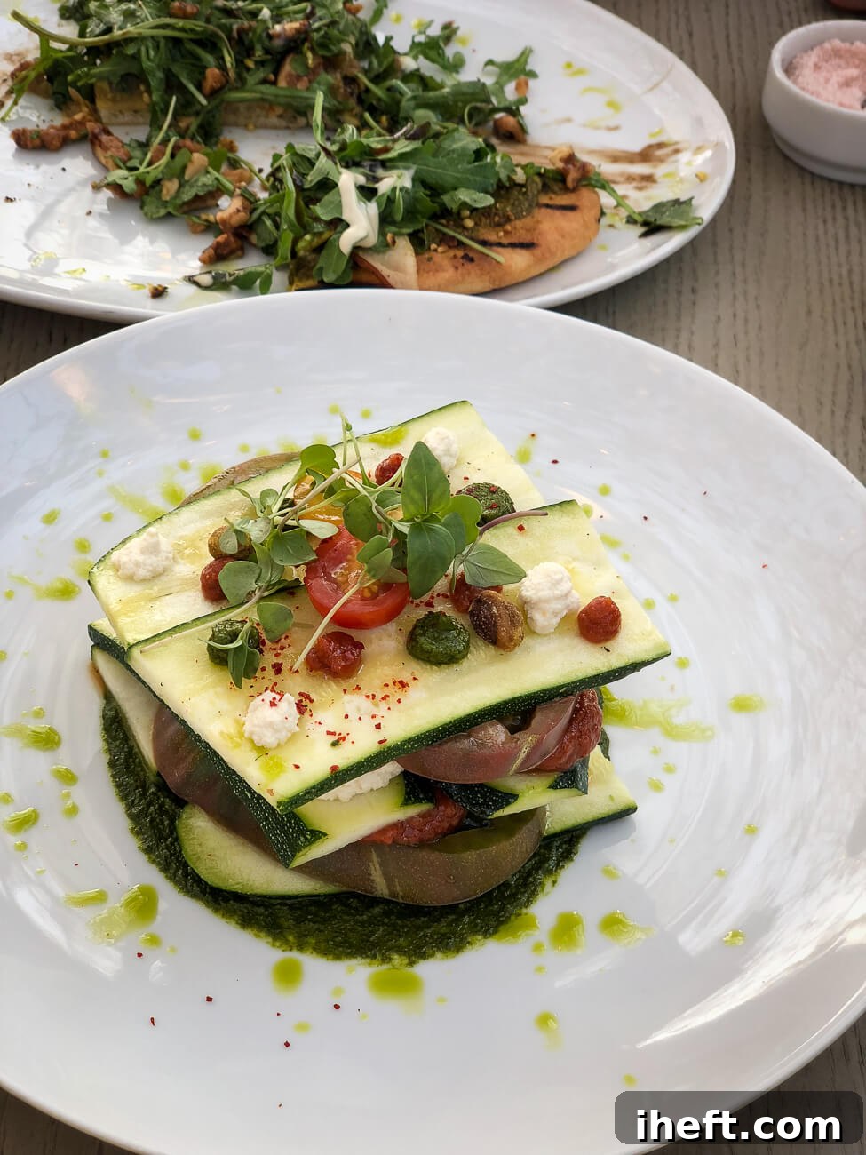 A pristine white plate featuring an elegant heirloom zucchini and tomato tower, beautifully layered and crowned with fresh microgreens, accompanied by rich tomato and creamy vegan ricotta sauces.