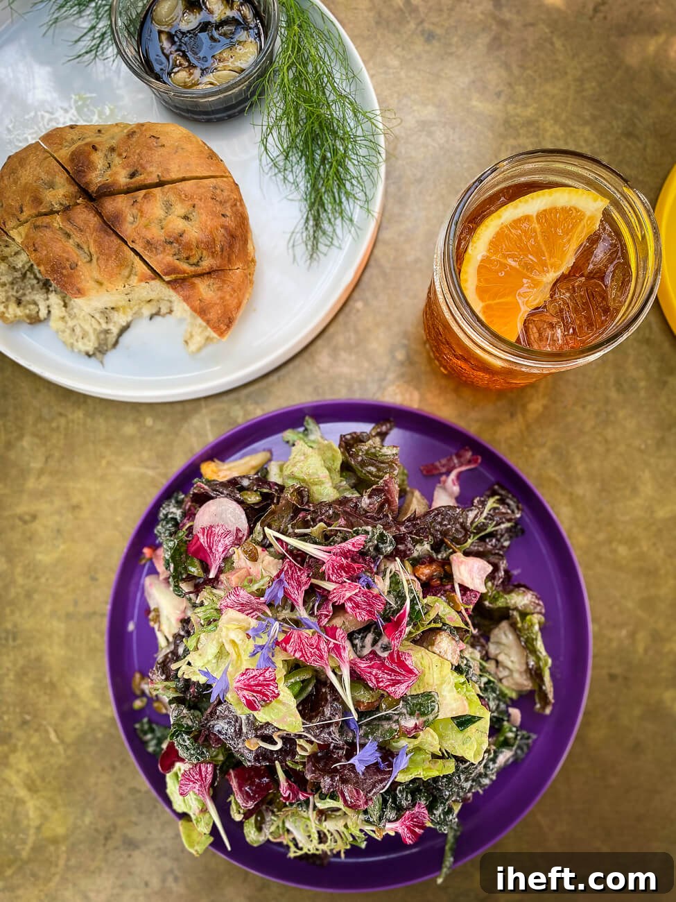 The signature "Yoga Pants" salad, beautifully plated on a purple dish, accompanied by a vibrant orange drink and a golden focaccia, at Satellite in Santa Barbara.