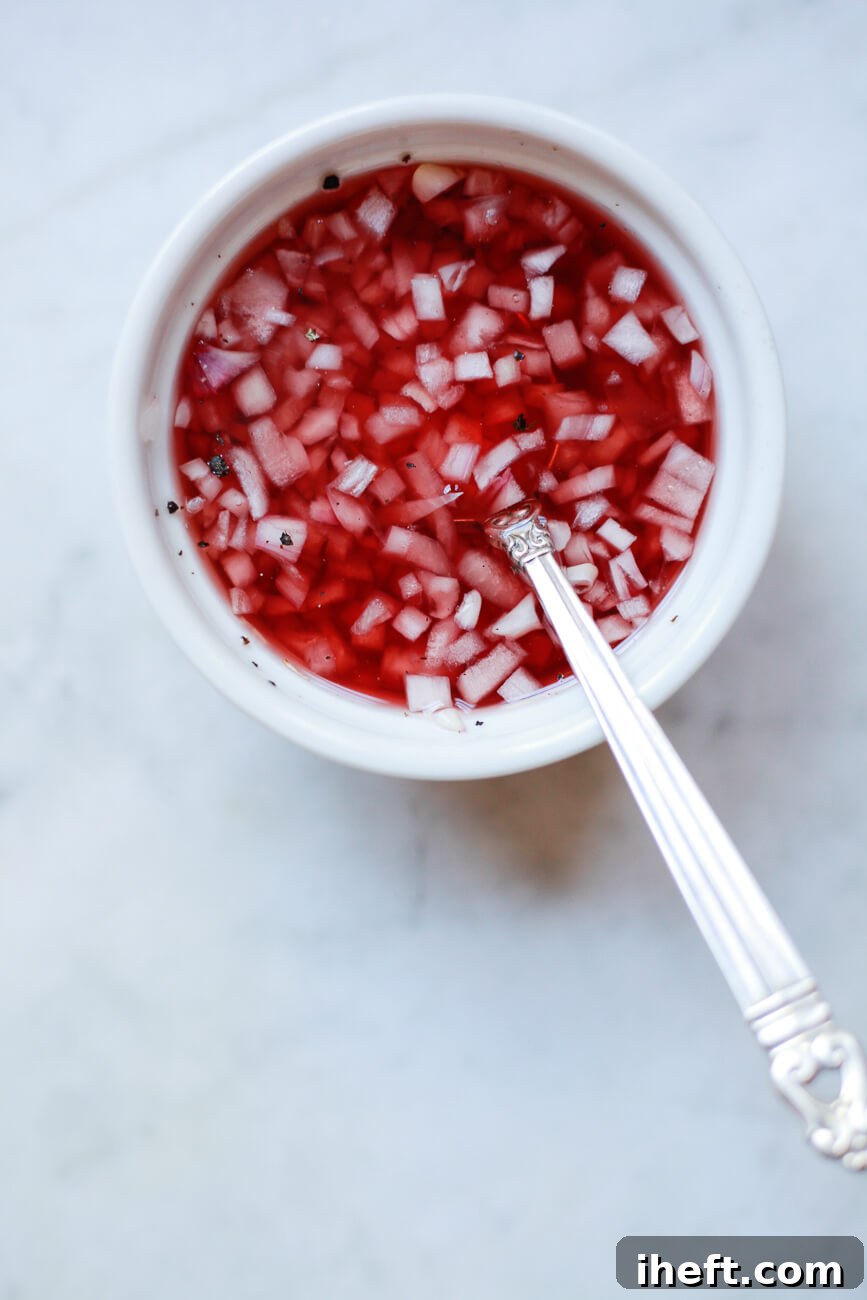 A small white bowl filled with red wine vinegar and shallot mignonette sauce for oysters. A small silver spoon sits in the bowl on a marble counter. 