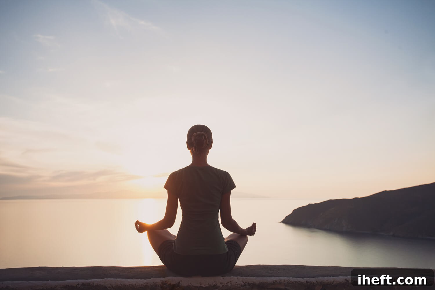 The back of a woman meditating sitting down cross-legged in front of a beautiful sunset over the ocean, symbolizing meditation and manifestation.