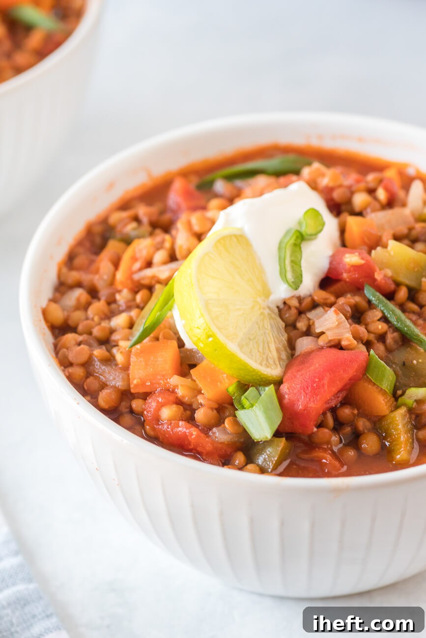 A close-up, mouth-watering photograph of delicious vegan lentil chili served in a pristine white ceramic bowl, elegantly placed on a matching white countertop. The chili is beautifully garnished with a wedge of fresh lime, vibrant green onions, and a dollop of creamy sour cream, highlighting its inviting appeal.
