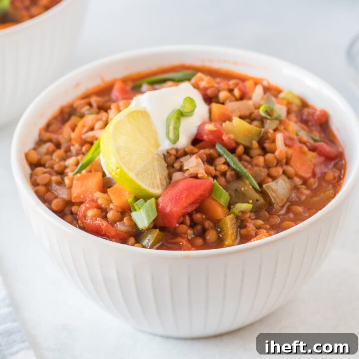 A beautifully presented bowl of lentil chili, garnished with fresh green onions, a dollop of sour cream, and a slice of lime, sitting on a table.