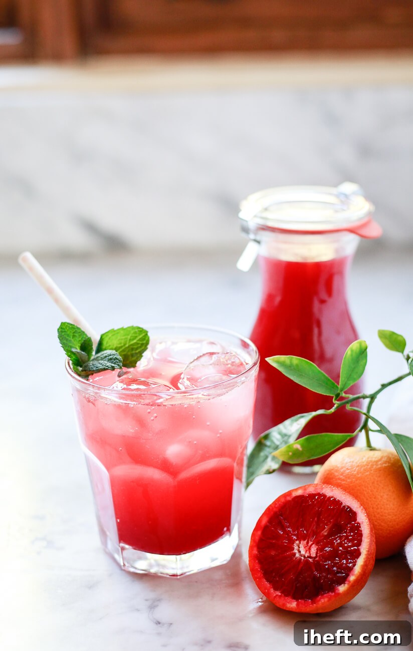 A short glass filled with sparkling water and blood orange juice garnished with mint and a paper straw. A small carafe of blood orange juice in the background and half a cut red orange in the foreground.