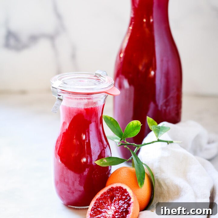 A small and large carafe filled with red blood orange juice on a marble countertop with a cut red orange in the foreground.