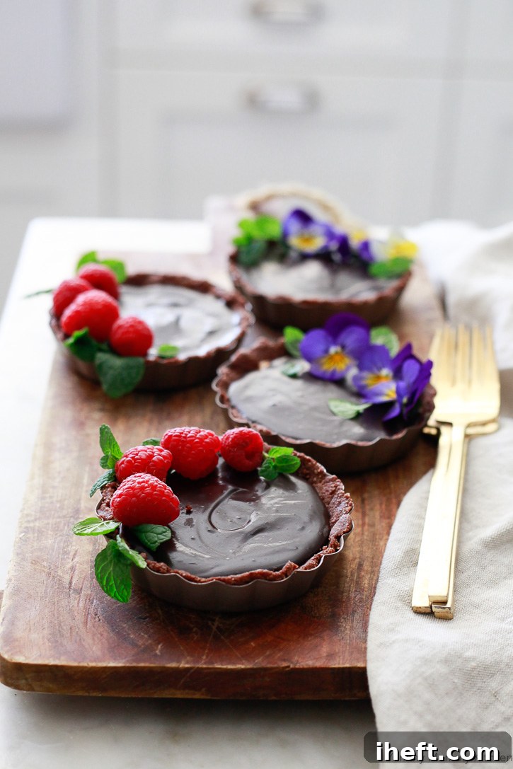 Beautiful no-bake chocolate almond butter tarts adorned with fresh raspberries and delicate edible flowers, showcasing their elegance and deliciousness.
