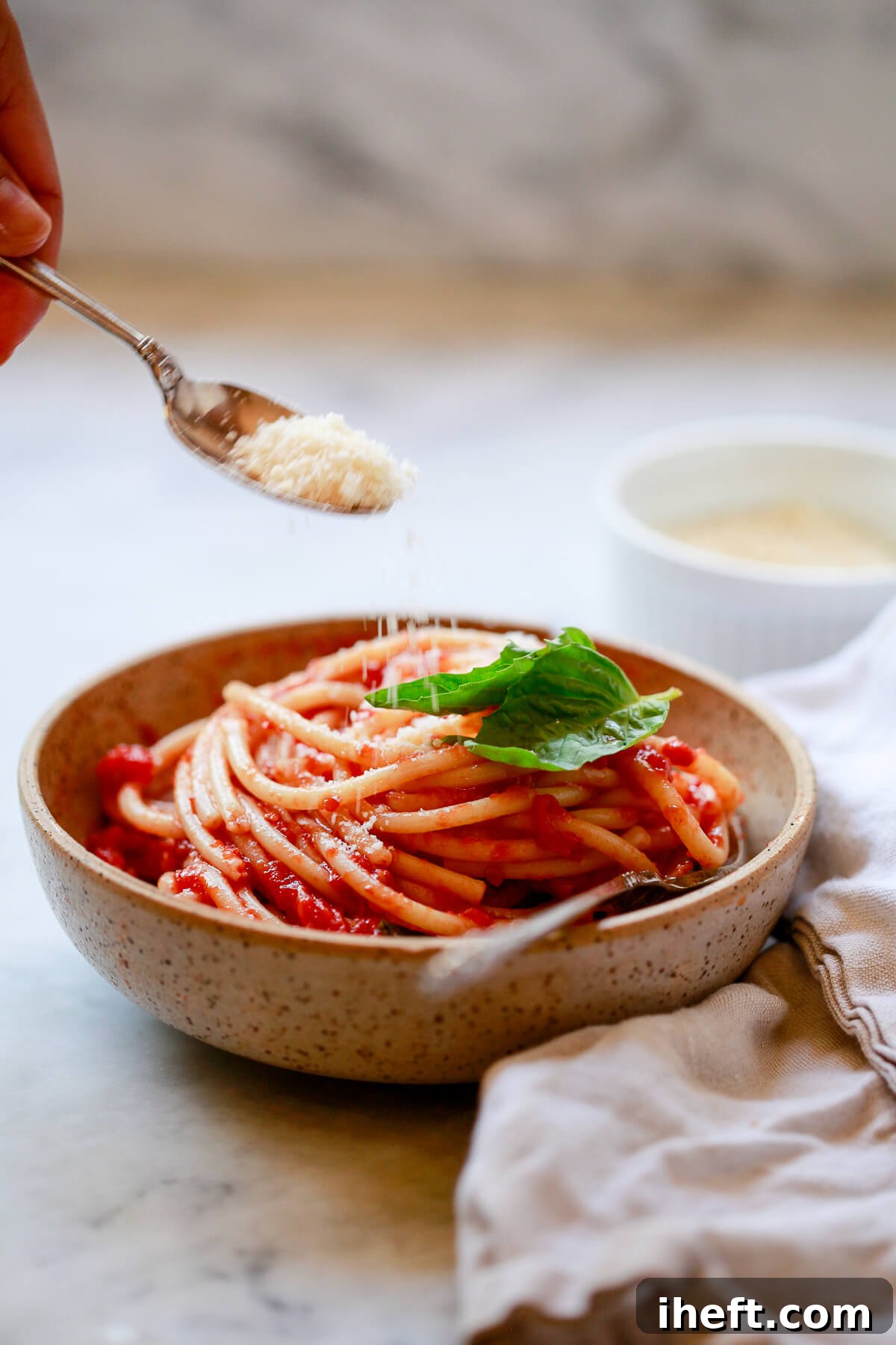A tan bowl brimming with spaghetti and homemade San Marzano marinara sauce is generously topped with grated Parmesan cheese, ready to be enjoyed.