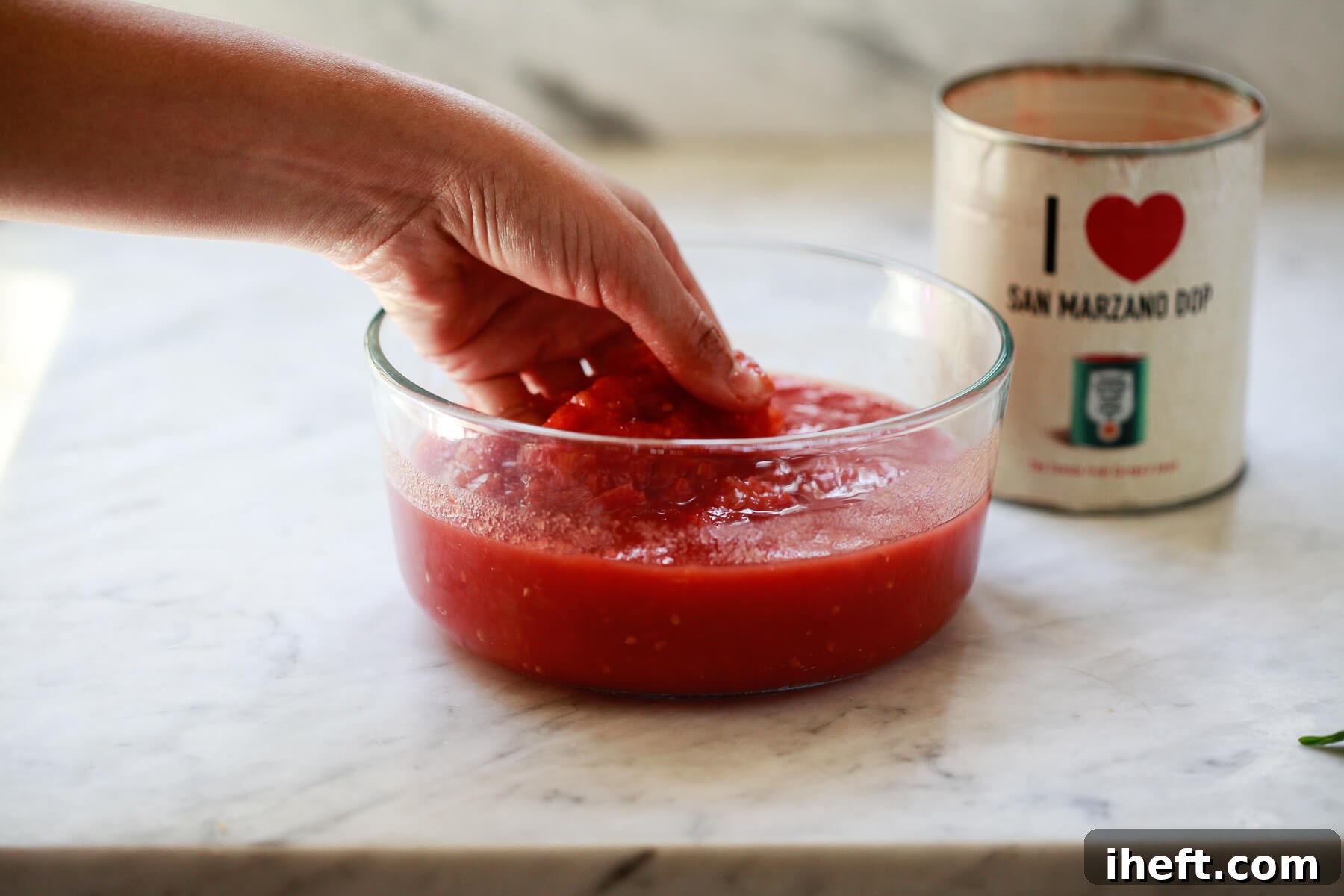 A hand crushing San Marzano tomatoes in a clear bowl.