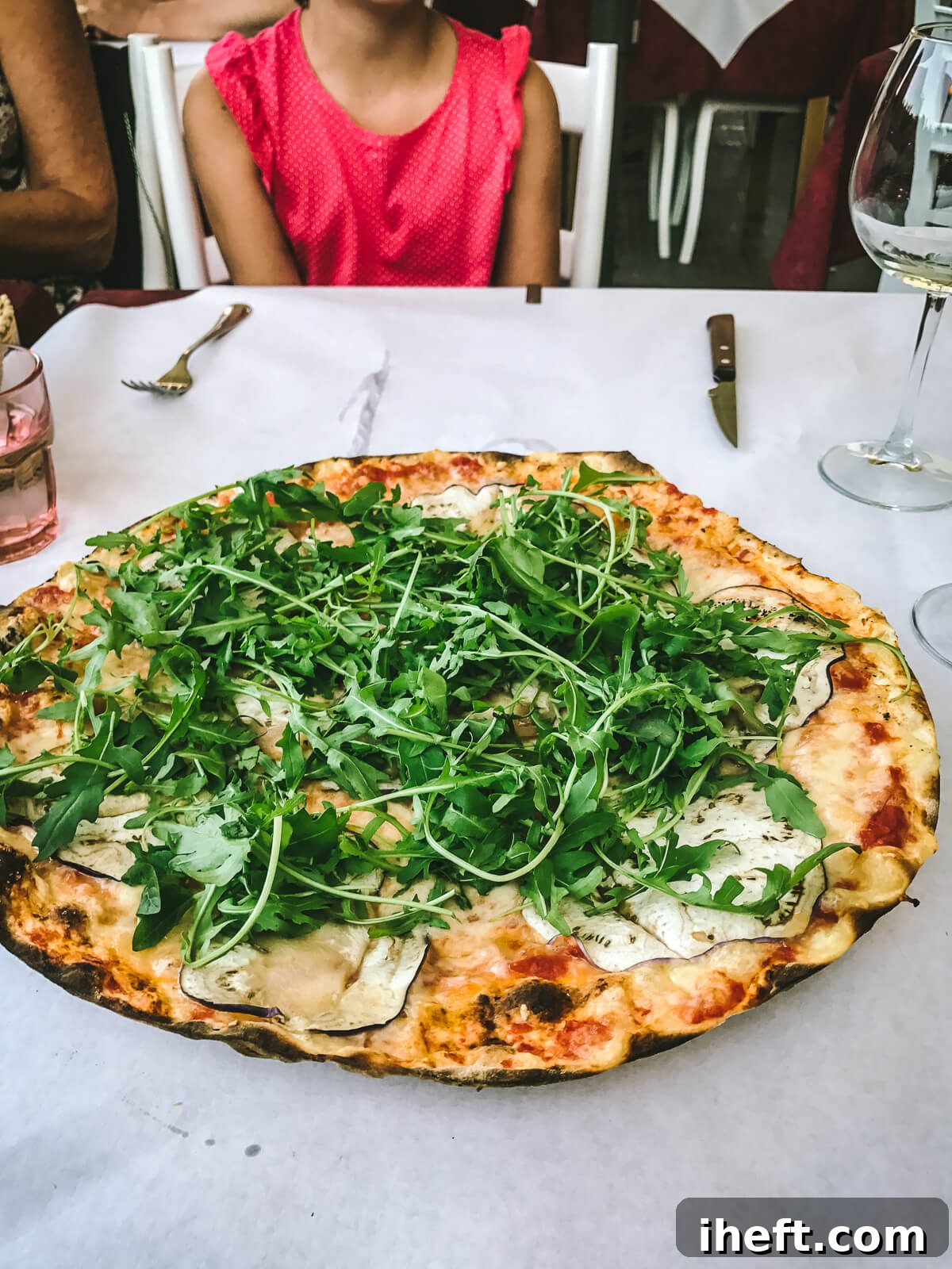 A large thin crust pizza with pizza sauce, cheese, eggplant and arugula on a white table in Tuscany Italy with a young girl in the background. 