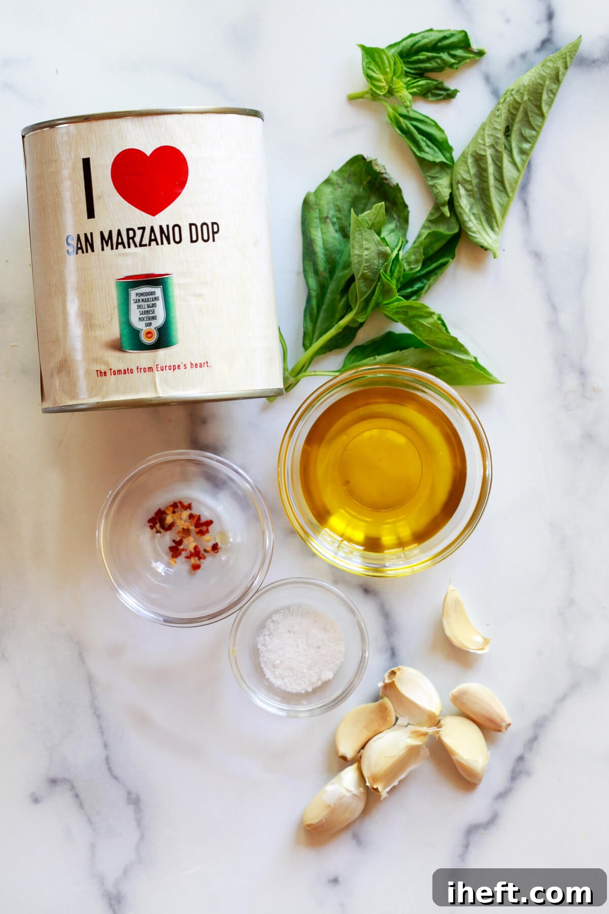 The ingredients for marinara sauce on a marble countertop: San Marzano DOP tomatoes, fresh basil, red pepper flakes, olive oil, salt, and garlic. 
