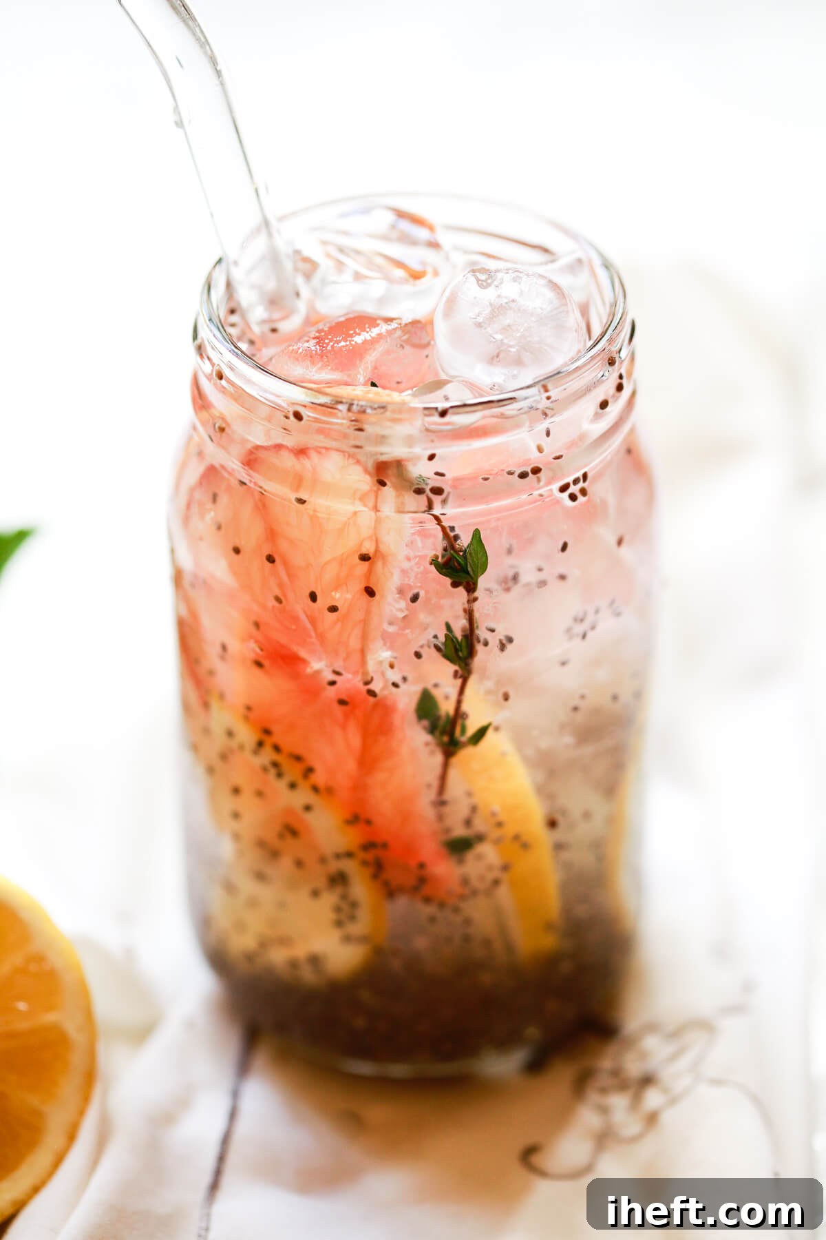 A mason jar filled with a chia drink made out of chia seeds in water, garnished with citrus slices and a sprig of fresh thyme, sits on a light surface.