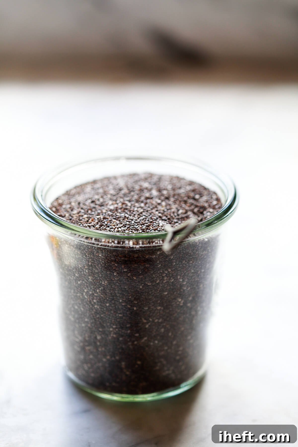 A glass Weck jar filled with dry chia seeds rests on a white marble countertop, accompanied by a small metal spoon.