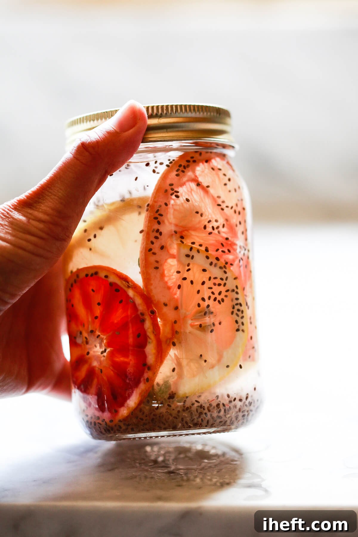 A mason jar filled with chia seeds in water and lemon slices is being picked up from a marble countertop, showcasing a beautiful detox drink.