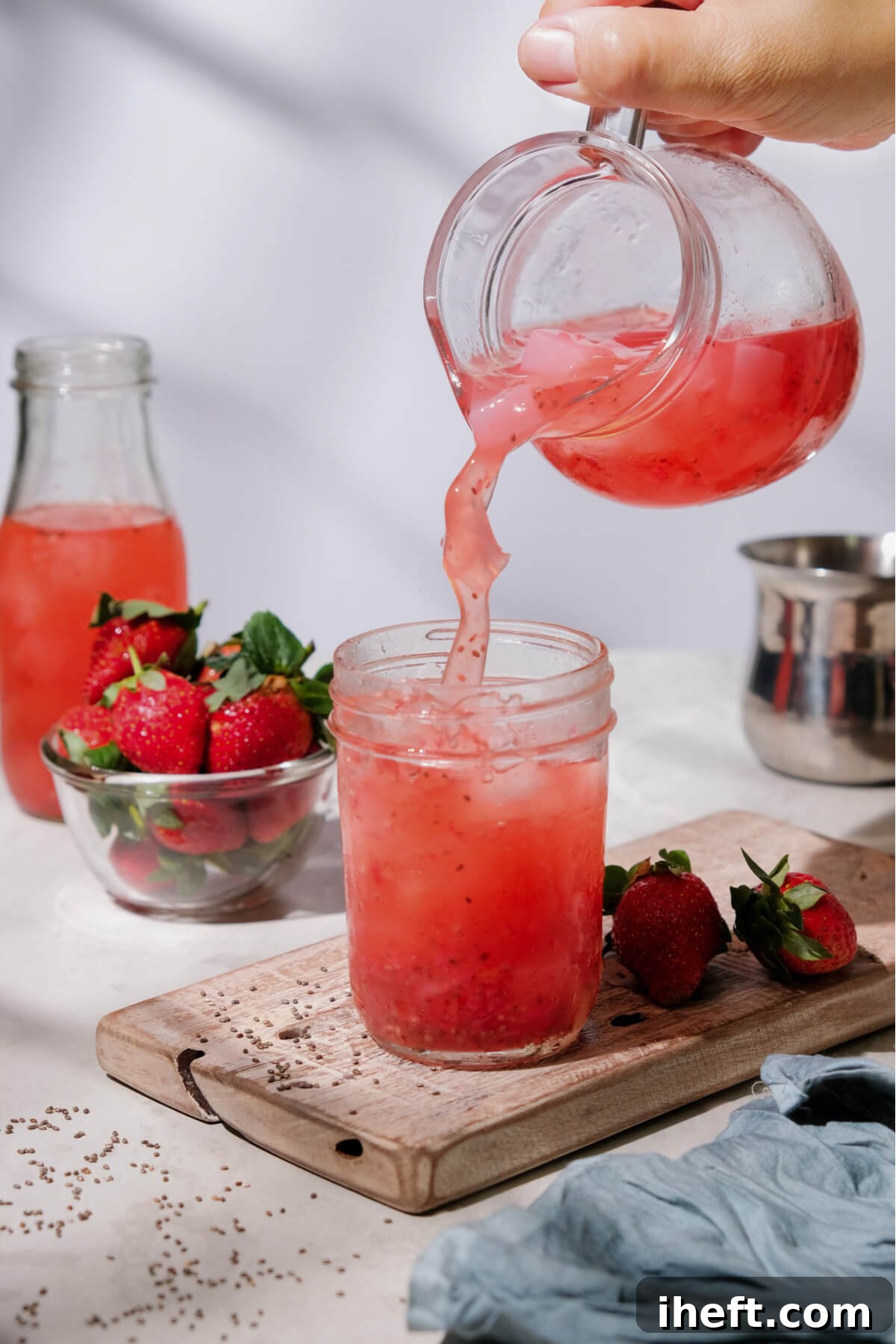 A glass pitcher pours a vibrant pink strawberry chia drink into a mason jar on a wooden board, ready to be enjoyed.